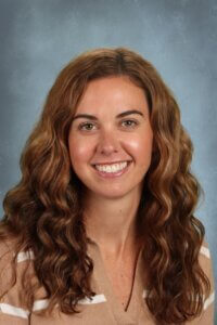 Smiling woman with long wavy brown hair in a beige striped sweater against a blue backdrop.