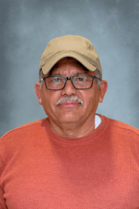 Portrait of a man wearing a beige cap, black-framed glasses, and an orange sweater, facing the camera against a blue backdrop.